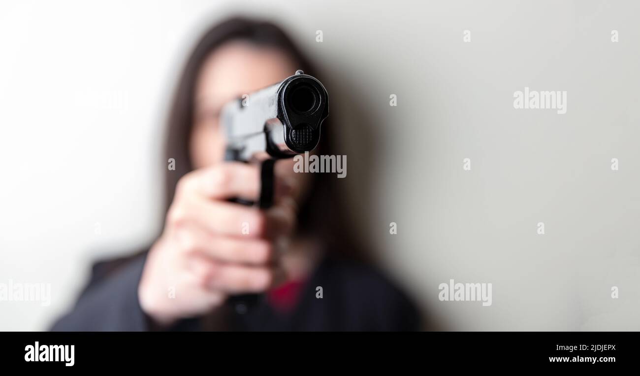 Woman holding a gun in hand pointing at camera. Female secret police agent, killer or spy dark long hair, copy space. Selective focus Stock Photo