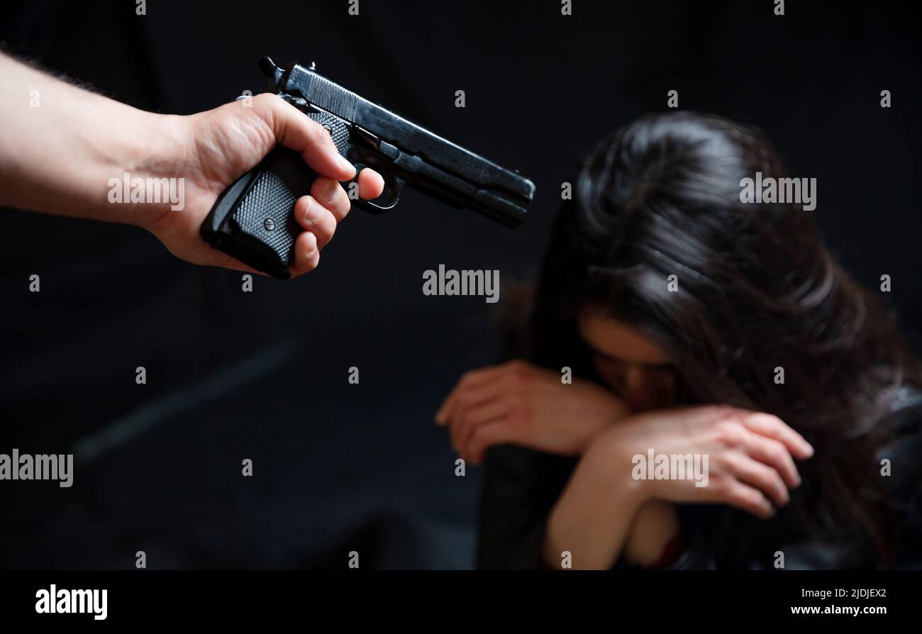 Threatened woman concept. Killer armed with a pistol terrorizes girl who keeps her head bent over. Criminal with gun, scared female, victim, violence, Stock Photo