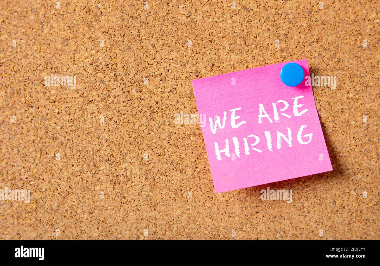 We Are Hiring phrase in pink post it on bulletin board cork background. Business vacancy text message, advertise, staff wanted announcement concept. C Stock Photo