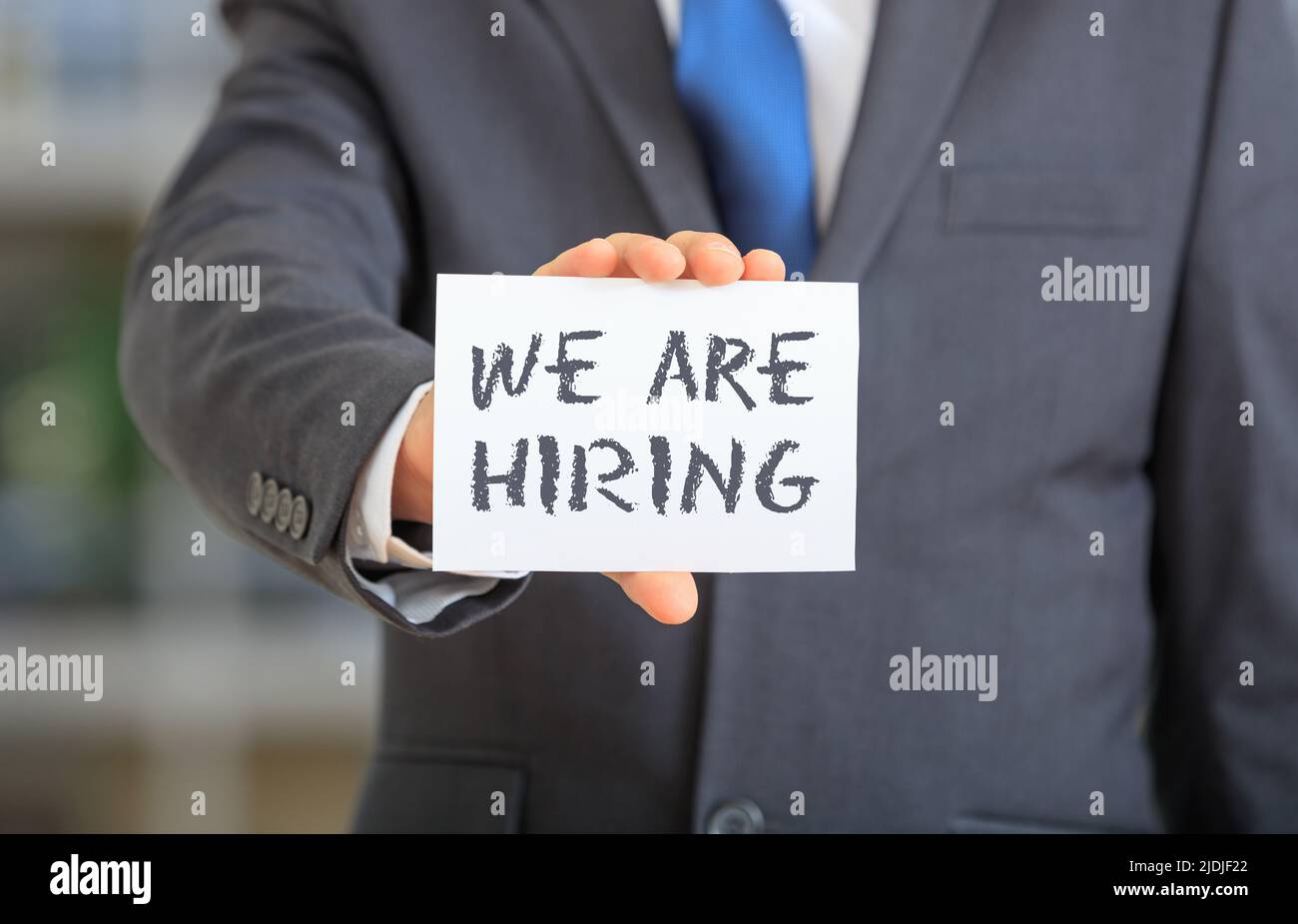 We Are Hiring white card in man hand. Man in blue suit holds a card with text message about business vacancy, advertise, staff wanted concept. Close u Stock Photo