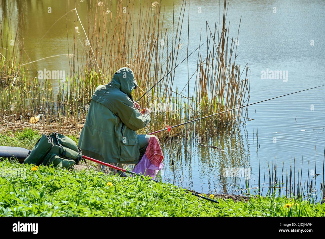 Riga, Latvia - May 8, 2022: A fisherman sits with a fishing rod on the shore of a lake in a public park. Green grass around. View from back. Stock Photo