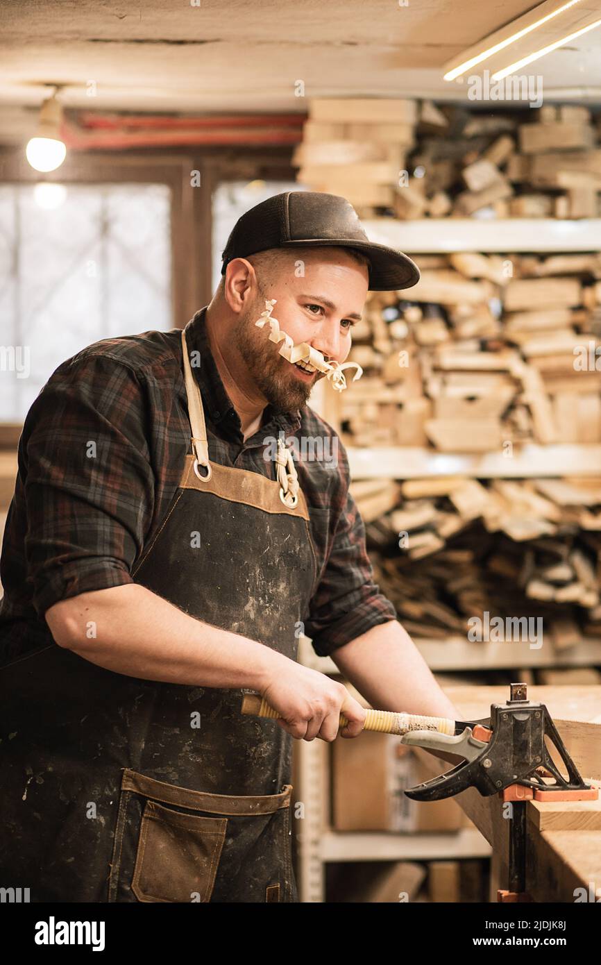Vertical playful man, carpenter in cap and protective apron with funny shaving chip mustache, learning to work with rasp Stock Photo