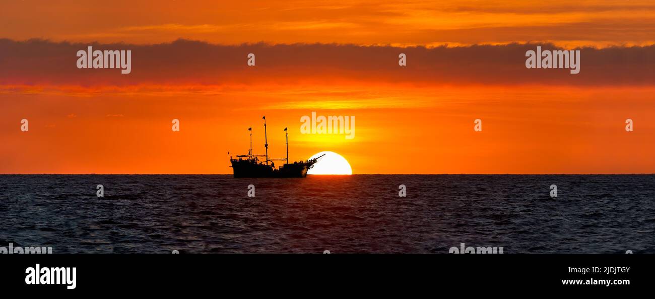A Silhouette Of A Ship Sits At Sea As The Sun Rises On the Ocean Horizon Stock Photo
