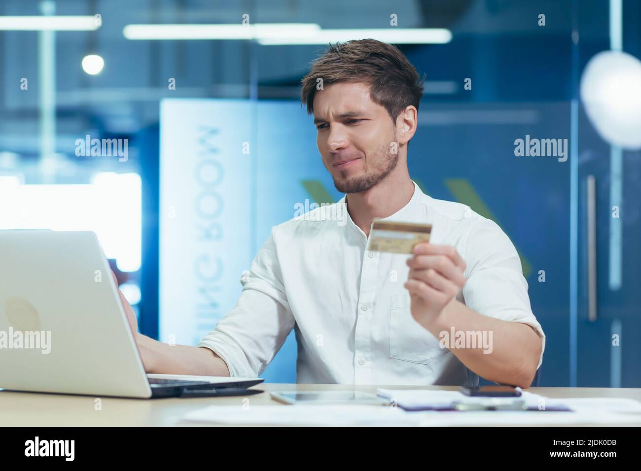 Upset and embarrassed young man, businessman. Holds a credit card in his hand, works on a laptop in the office. Pays loans, bills Stock Photo