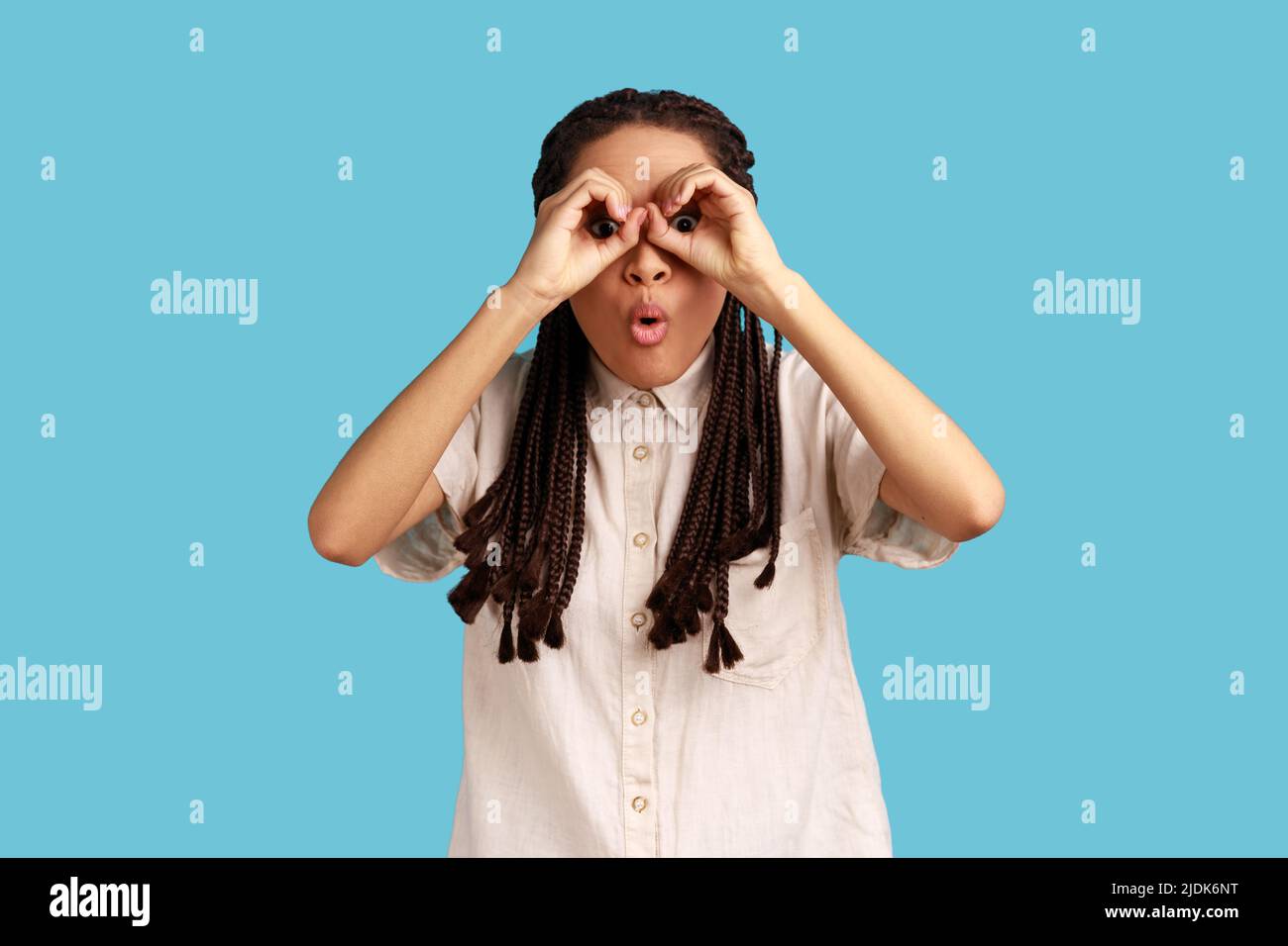 Funny playful woman with black dreadlocks covering eyes with ok signs, makes binoculars, foolishes around and looks through goggles, wearing white shirt. Indoor studio shot isolated on blue background Stock Photo