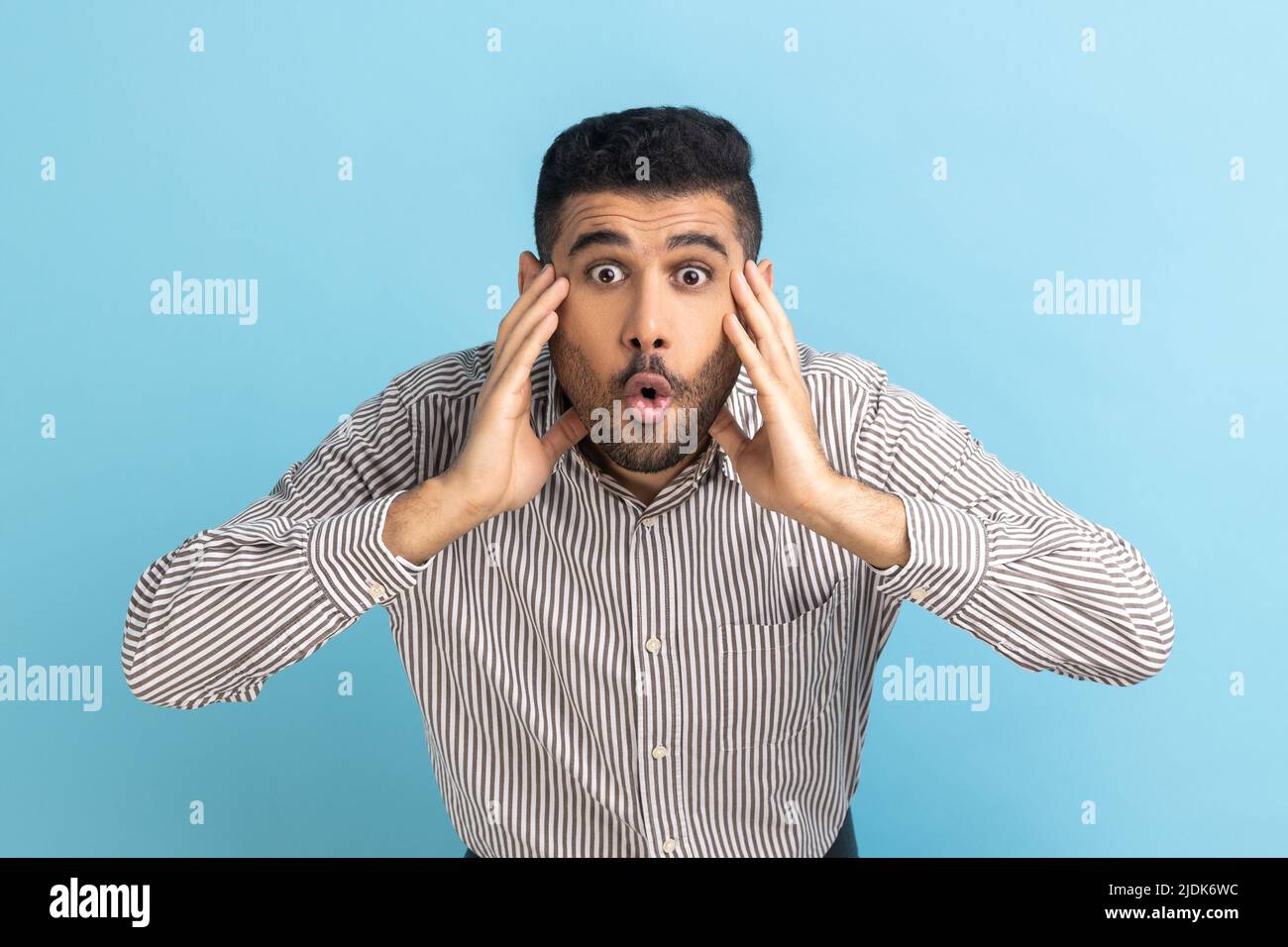 Wow, it's unbelievable. Surprised businessman staring at camera with widely open mouth and big eyes, raising arms in amazement, wearing striped shirt. Indoor studio shot isolated on blue background. Stock Photo