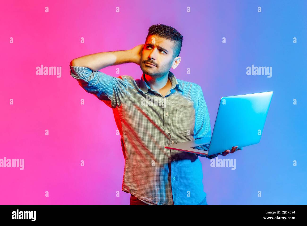 Portrait of handsome pensive man in shirt holding laptop and rubbing head looking away, thinking over new business project. Indoor studio shot isolated on colorful neon light background. Stock Photo