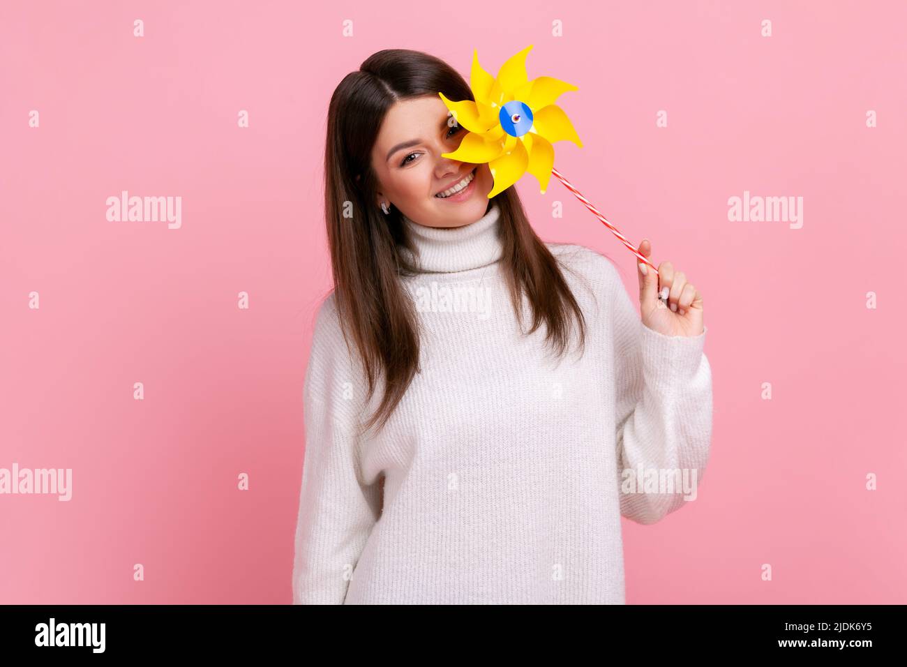 Childish dark haired female covering eye with paper windmill, playing with pinwheel toy on stick, wearing white casual style sweater. Indoor studio shot isolated on pink background. Stock Photo