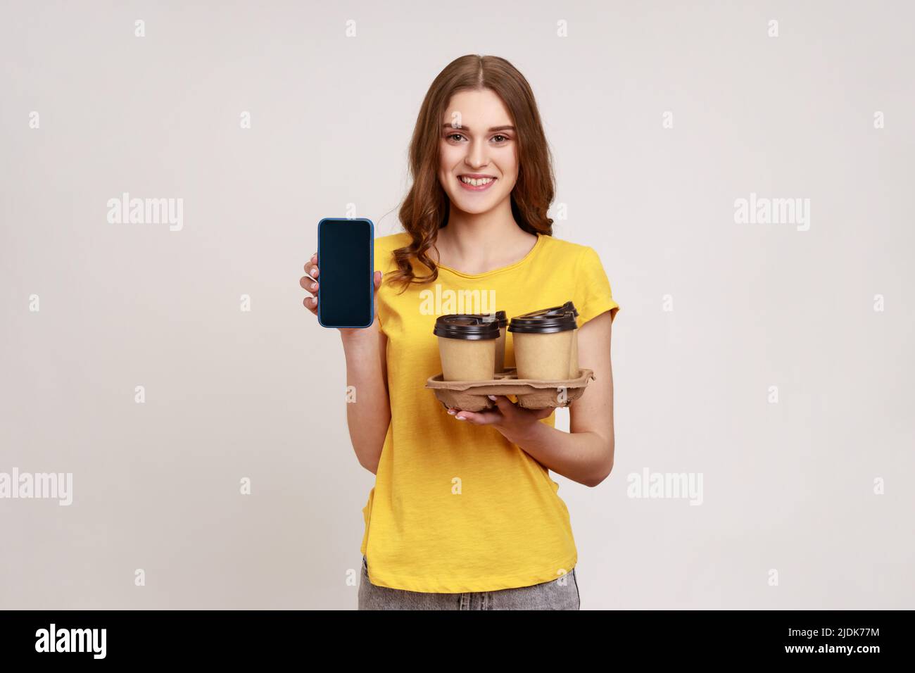 Positive smiling teenager girl with brown hair in yellow t-shirt holding empty display smartphone and holder with coffee paper cups, online order. Indoor studio shot isolated on gray background. Stock Photo