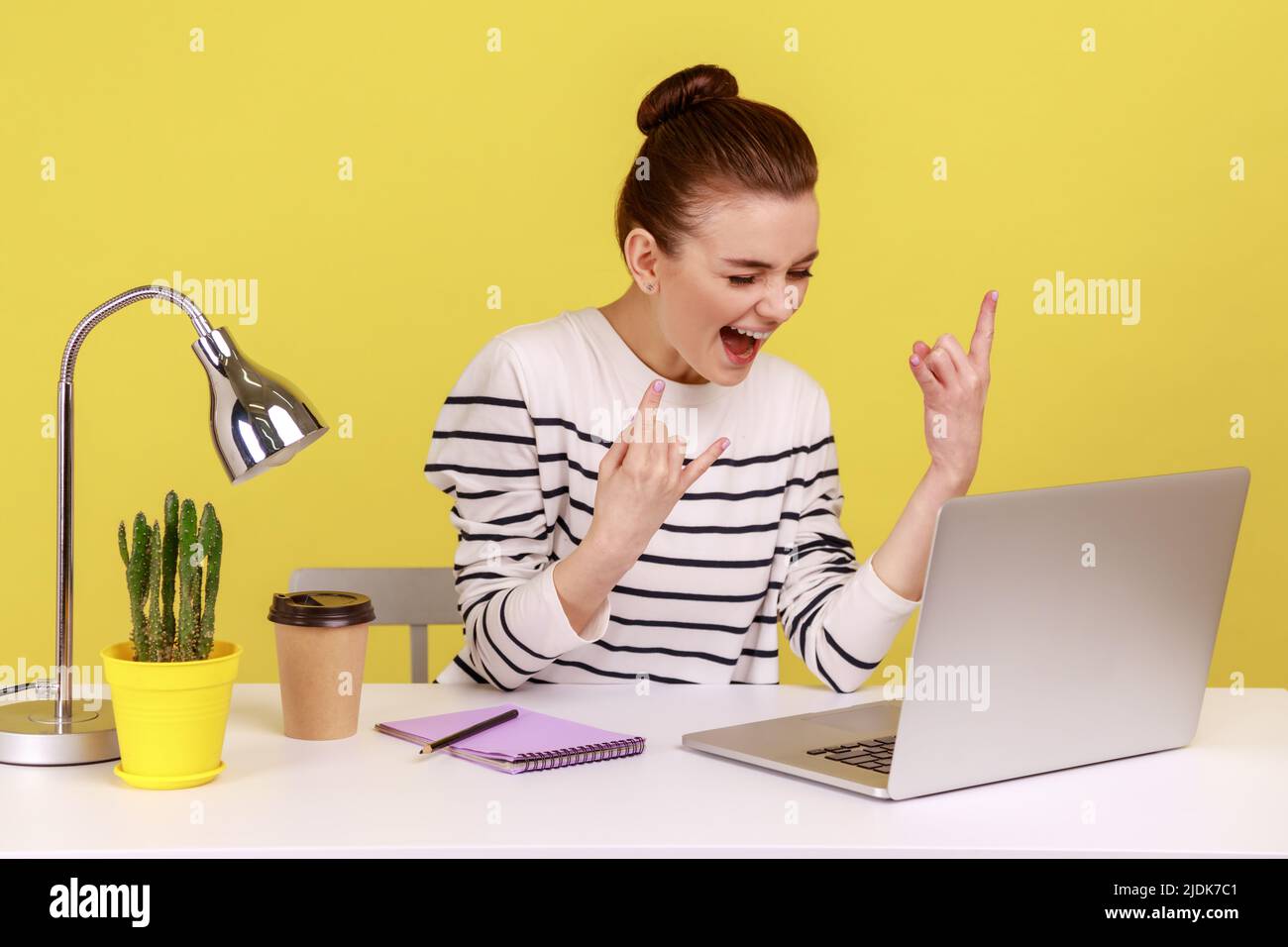 Excited naughty woman blogger showing rock and roll finger gesture looking at laptop screen, chatting web camera sitting at workplace. Indoor studio studio shot isolated on yellow background. Stock Photo