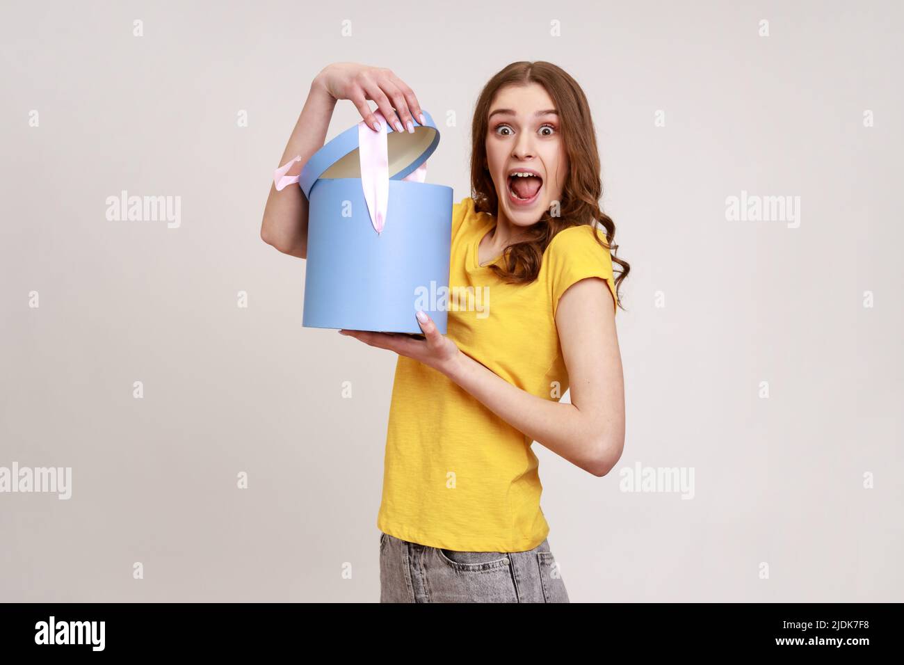 Amazed teenager girl in yellow casual T-shirt opening gift box, having excited surprised expression, unpacking birthday present surprise. Indoor studio shot isolated on gray background. Stock Photo