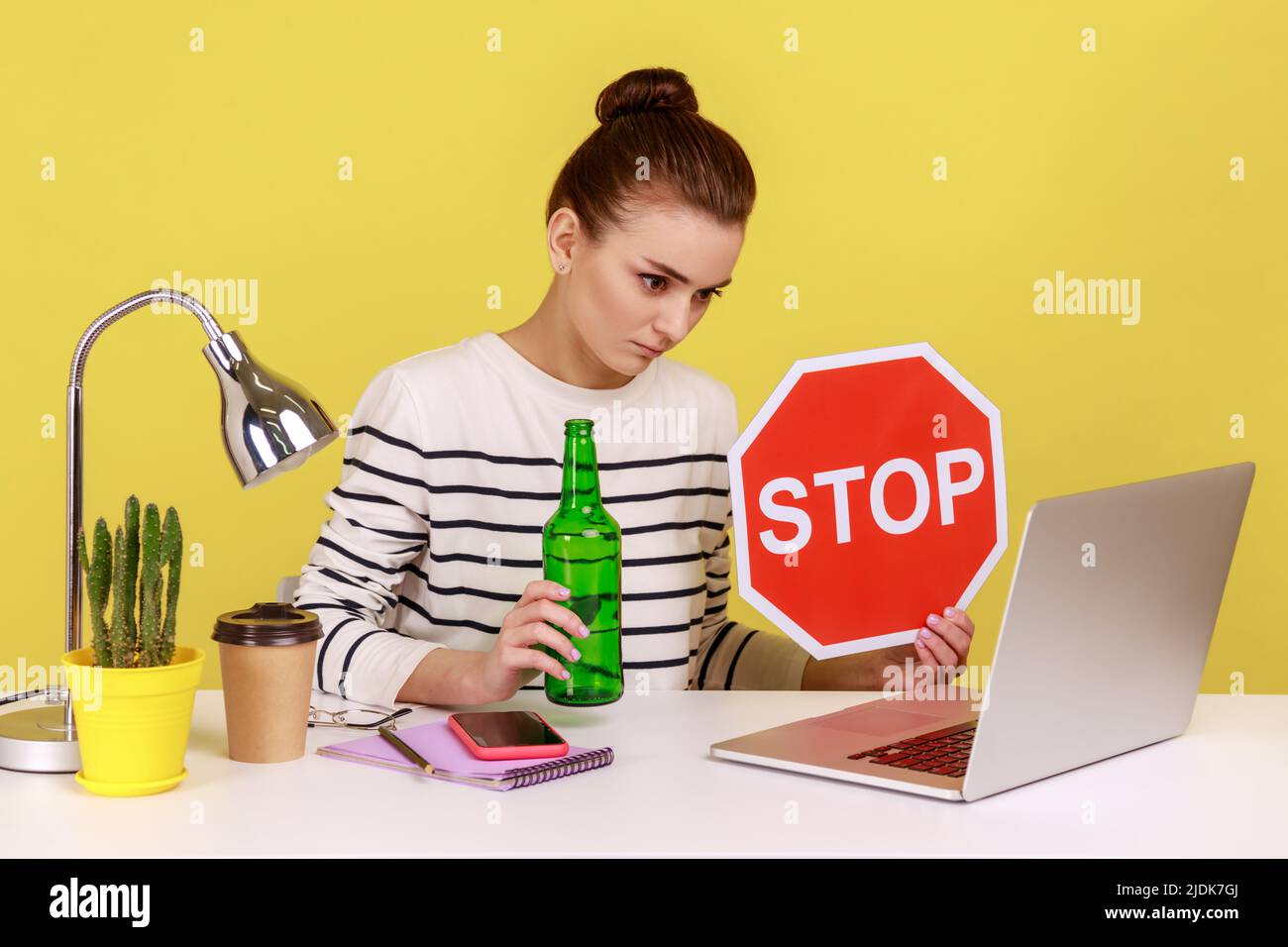Woman office worker in striped shirt sitting on workplace and having video conference, showing alcohol bottle and red stop sign, Indoor studio studio shot isolated on yellow background. Stock Photo