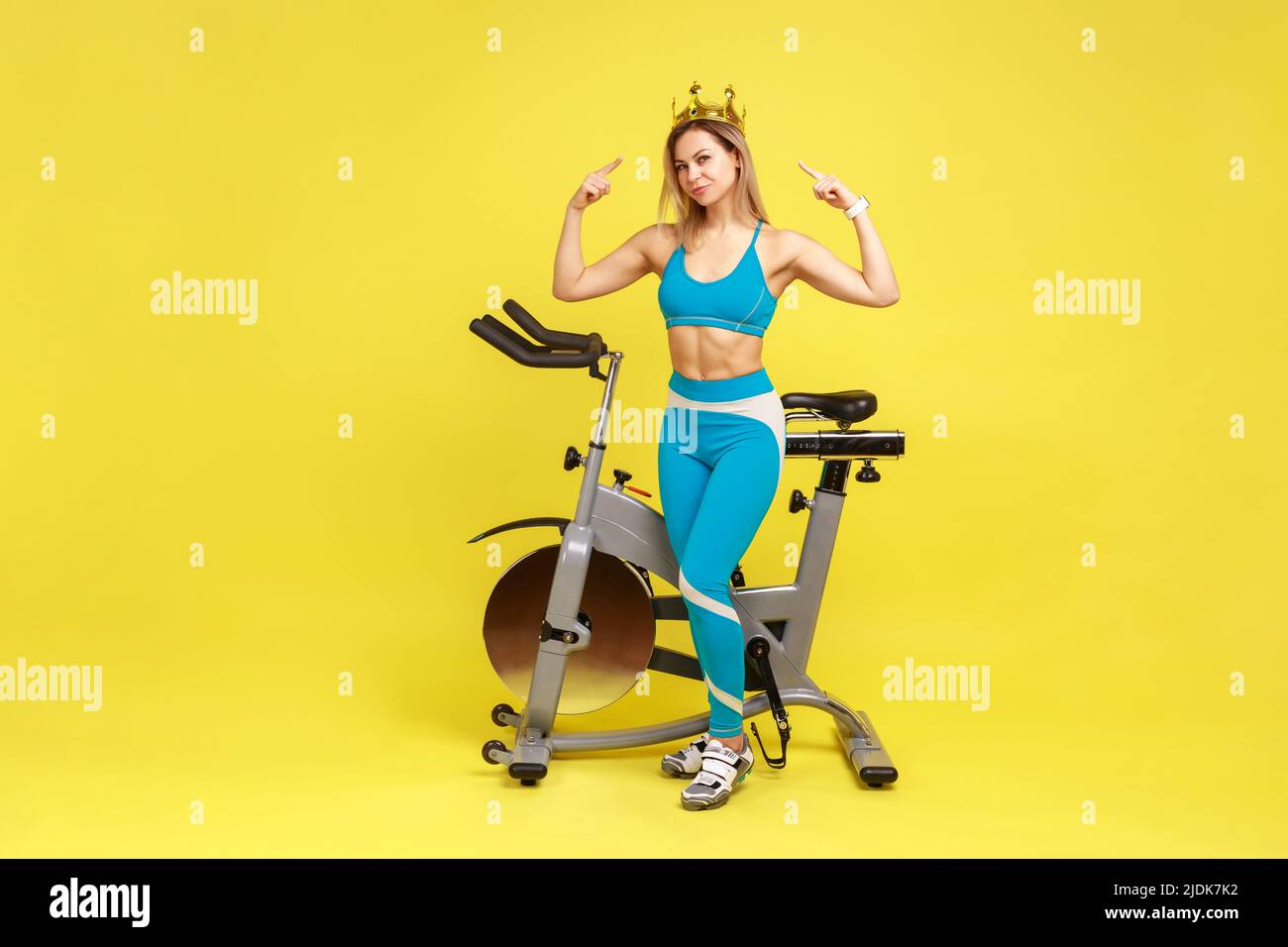 Full length portrait of woman sport coach standing pointing at golden crown on her head, being queen of fitness, wearing blue sportswear. Indoor studio shot isolated on yellow background. Stock Photo