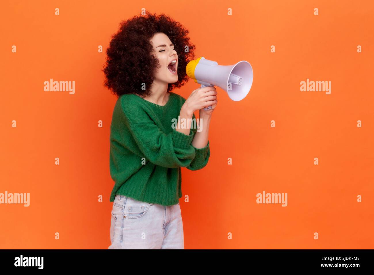 Side view portrait of woman with Afro hairstyle wearing green casual style sweater protesting or making announcement, screaming in megaphone. Indoor studio shot isolated on orange background. Stock Photo