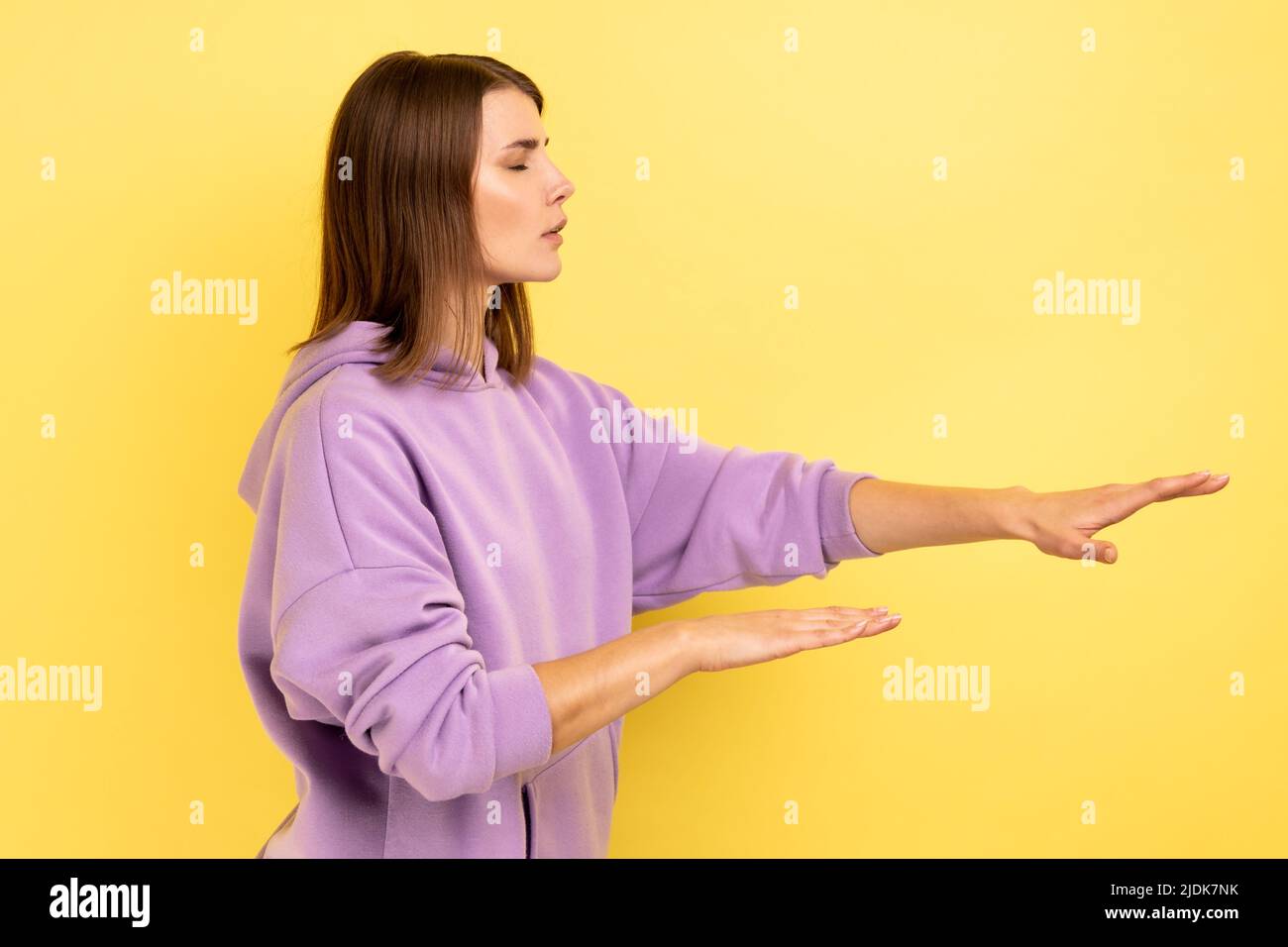 Side view of woman with eyesight problems trying to move with closed eyes touching space around with hands, vision disability, wearing purple hoodie. Indoor studio shot isolated on yellow background. Stock Photo