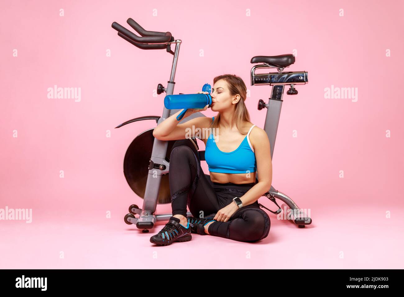 Portrait of thirsty athletic woman drinking water from bottle, restoring water balance after exhausted workout, wearing sports tights and top. Indoor studio shot isolated on pink background. Stock Photo