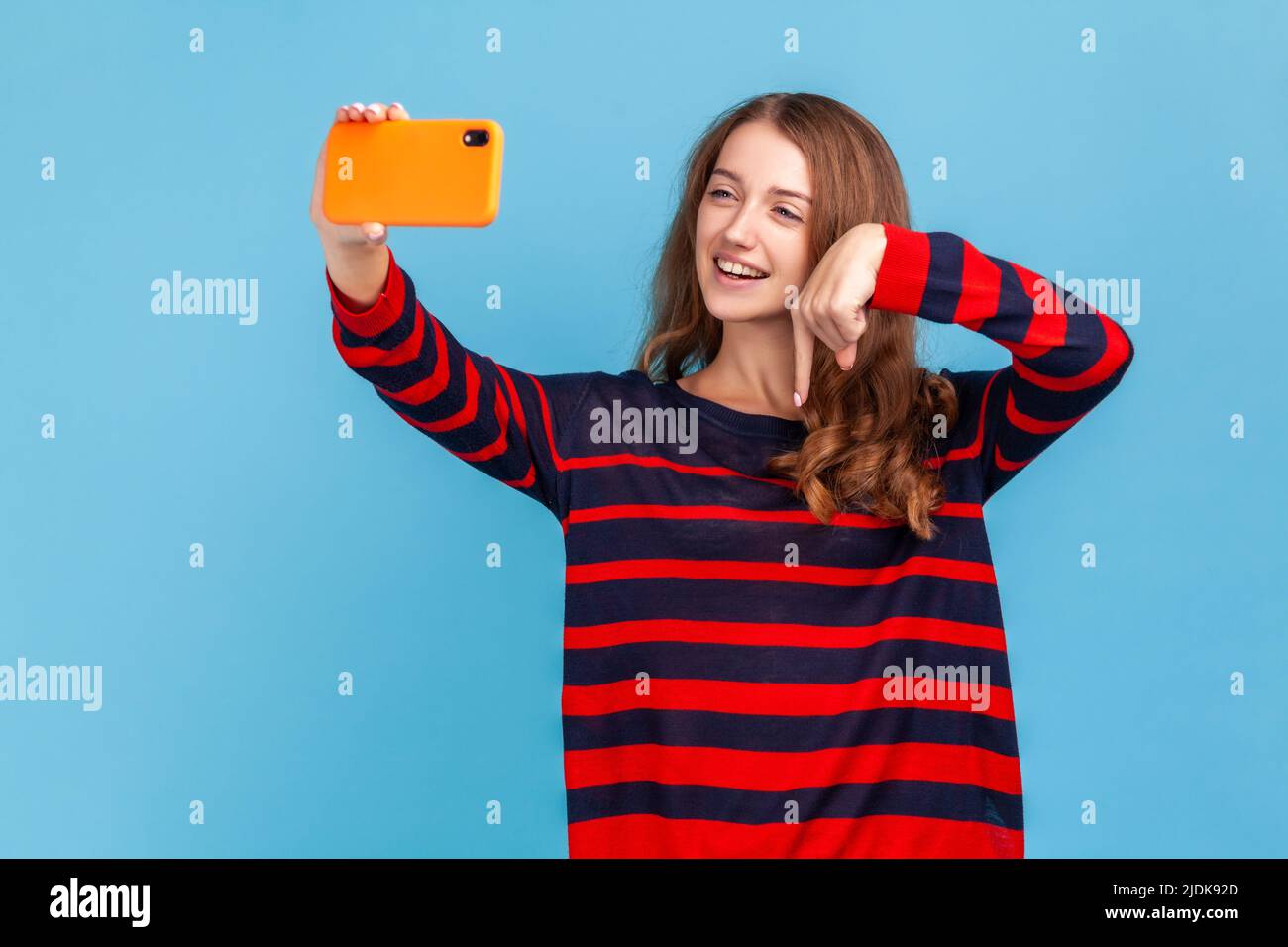 Attractive woman wearing striped casual style sweater, broadcasting livestream, pointing finger down, asking to subscribe for her blog. Indoor studio shot isolated on blue background. Stock Photo