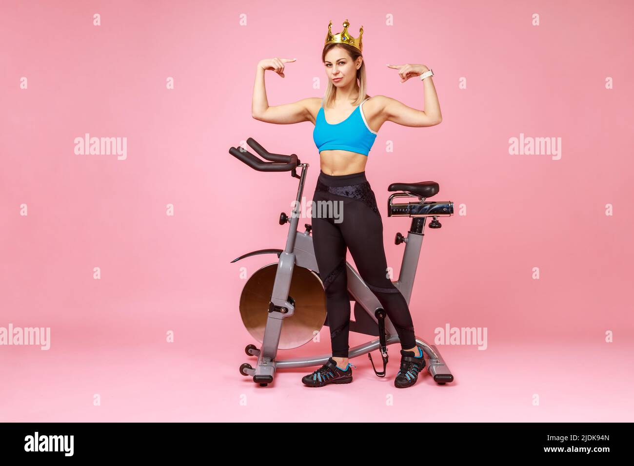 Full length portrait of slim confident sporty woman in golden crown standing near exercise bike and and showing biceps, raising arms. Indoor studio shot isolated on pink background. Stock Photo