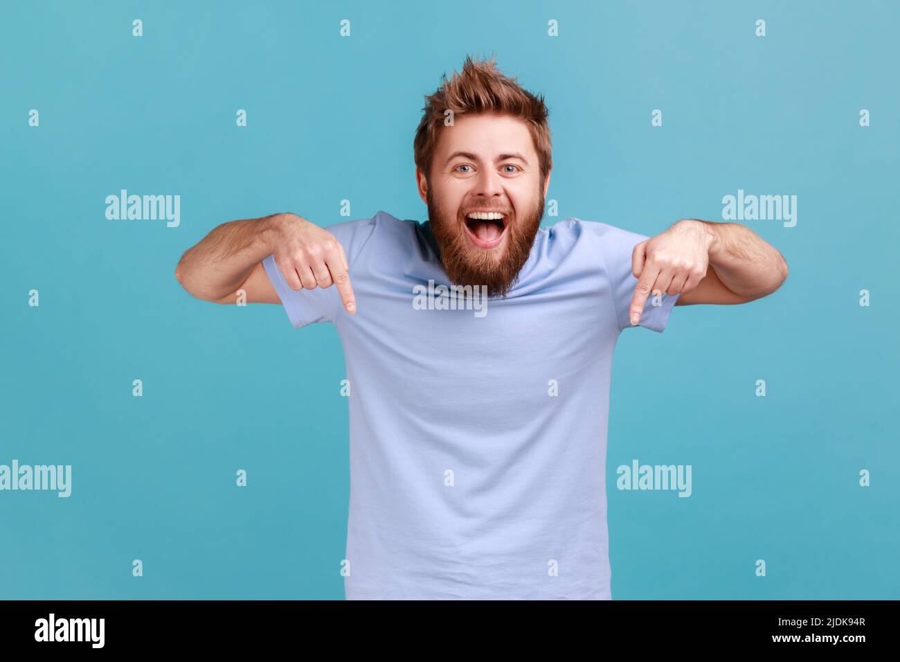 Portrait of happy satisfied bearded man pointing down and looking at camera, showing place for idea presentation, expressing positive emotions. Indoor studio shot isolated on blue background. Stock Photo
