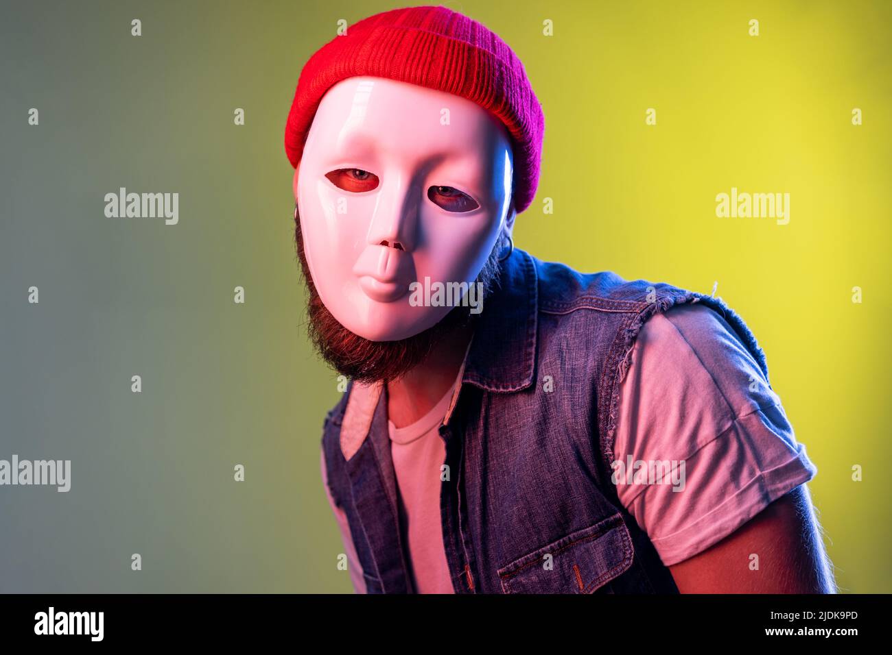 Portrait hipster man in white mask, spying undercover, corruption, hiding his personality, wearing beanie hat and denim vest. Indoor studio shot isolated on colorful neon light background. Stock Photo
