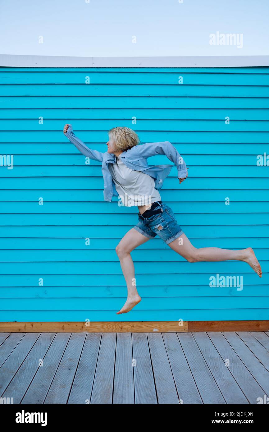 Side view of middle-aged woman superwoman with short fair hair flying like superhero, hurrying up at blue wooden wall. Stock Photo