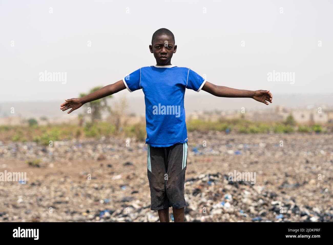 Desperate little African boy standing with arms outstretched in the middle of a desert area covered with dry plants and lots of garbage;symbol of poll Stock Photo