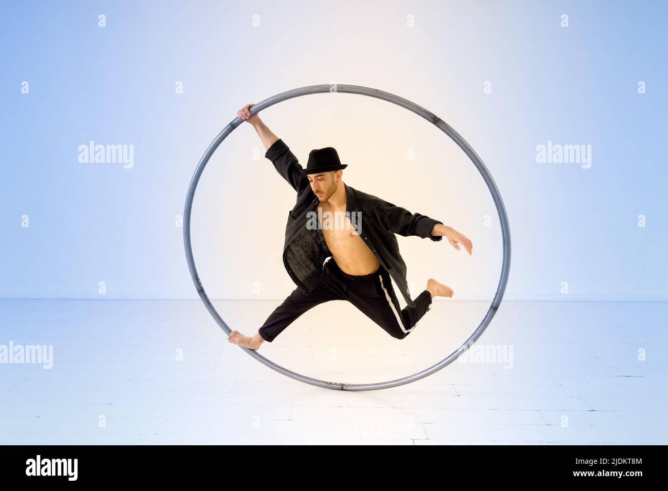 Full body of active male acrobat performing trick with cyr wheel while standing on one leg with raised arm against light background Stock Photo