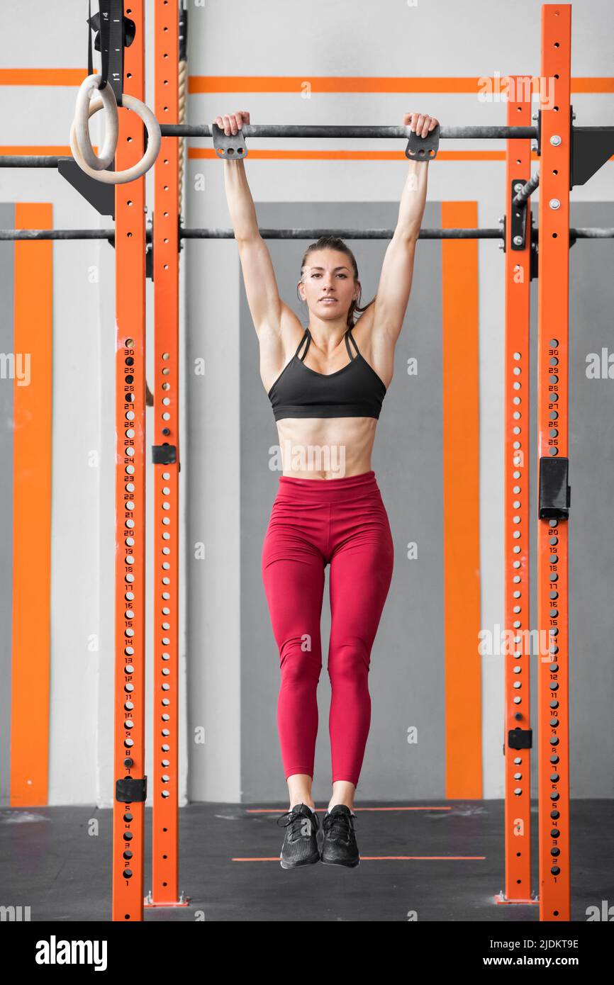 Fit athletic woman doing a dead hang from bars in a Crossfit gym in a full length view for a healthy active lifestyle concept Stock Photo