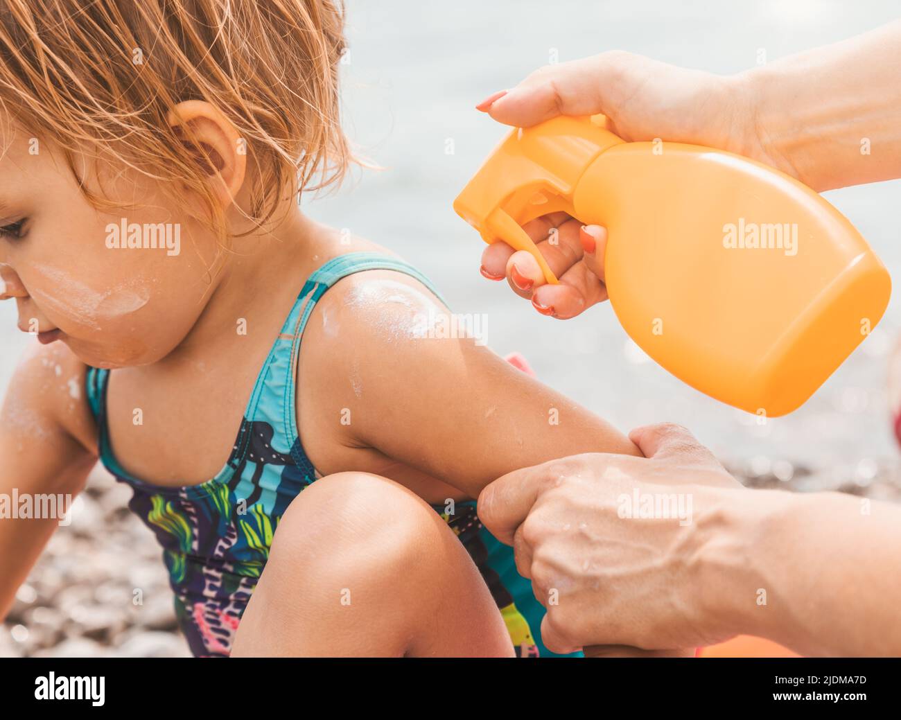 Mother spraying sunblock cream on her daughter's skin before sunbathing on a beach Stock Photo