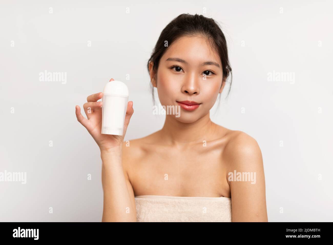Young chinese lady wrapped in towel showing deodorant, posing after shower on white studio background Stock Photo