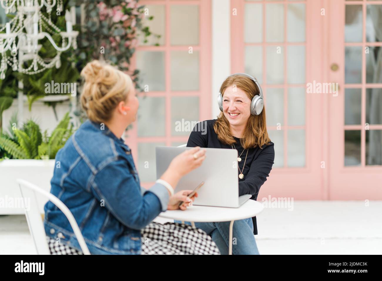 Two smiling women sitting at table with laptop and phone wearing headphones Stock Photo