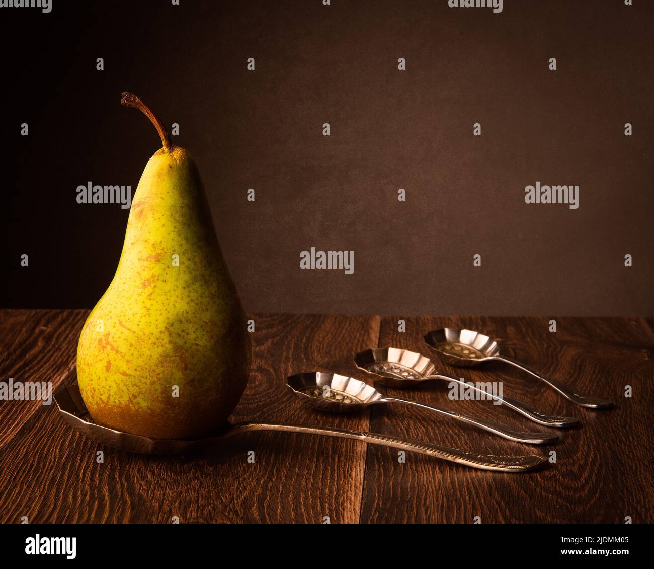 Still life of a pear on a silver spoon along side three more spoons on a dark wood table with a dark background for copy Stock Photo