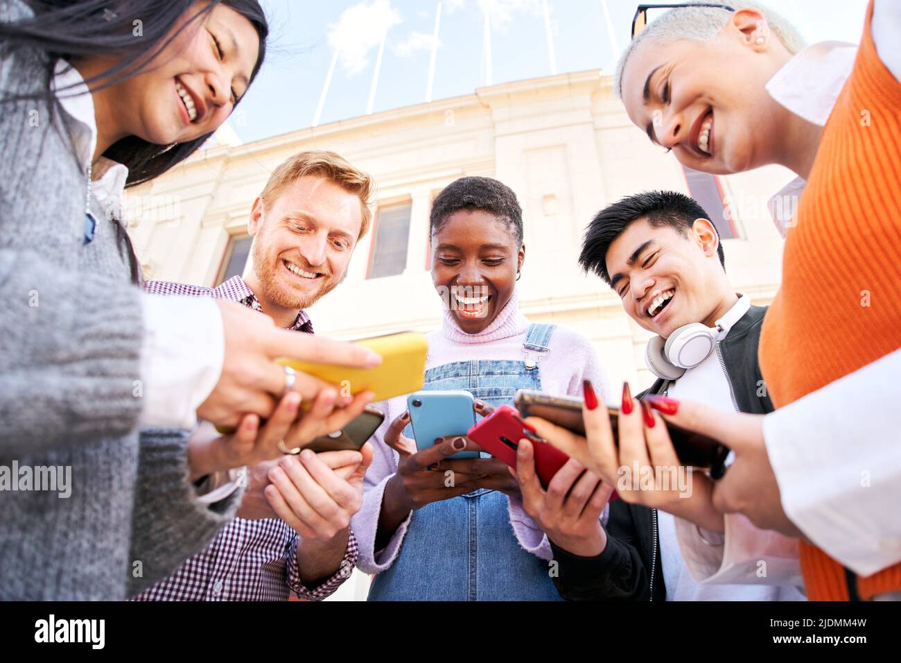 Low angle Group of Smiling people in circle using mobile outdoors. Cheerful friends having fun watching something at smart phone. Happy Social media Stock Photo