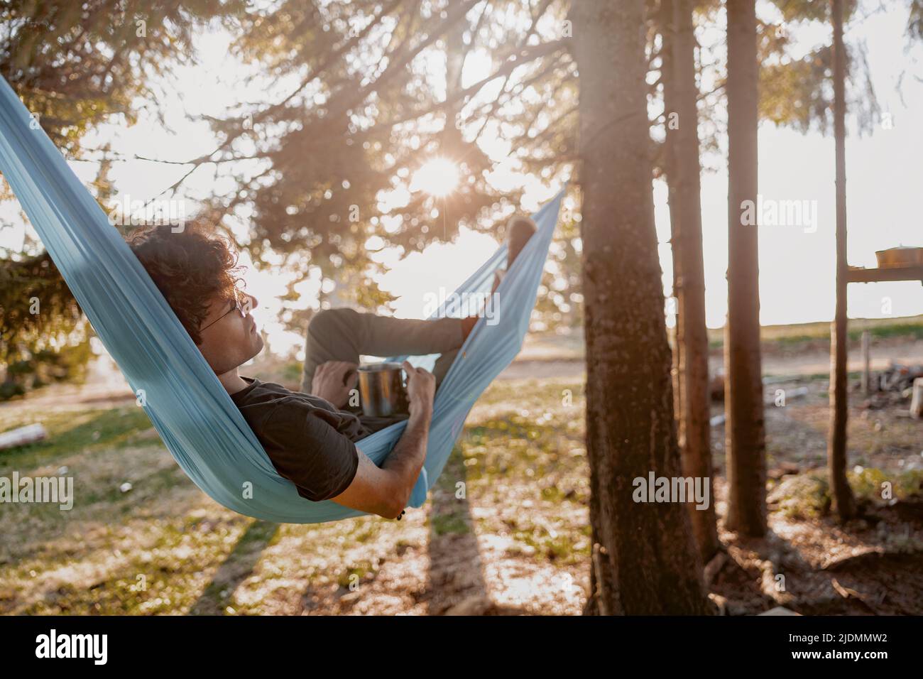 Attractive young man resting in hammock with coffee, tea in hammock in forest. Rear. Stock Photo