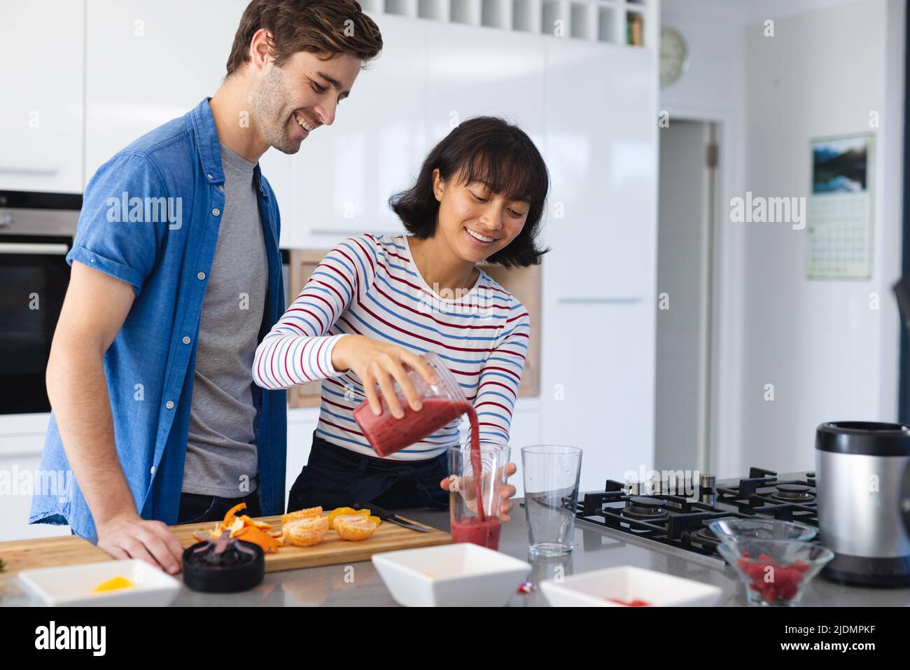 Smiling caucasian boyfriend looking at asian girlfriend pouring fruits smoothie in glass in kitchen Stock Photo