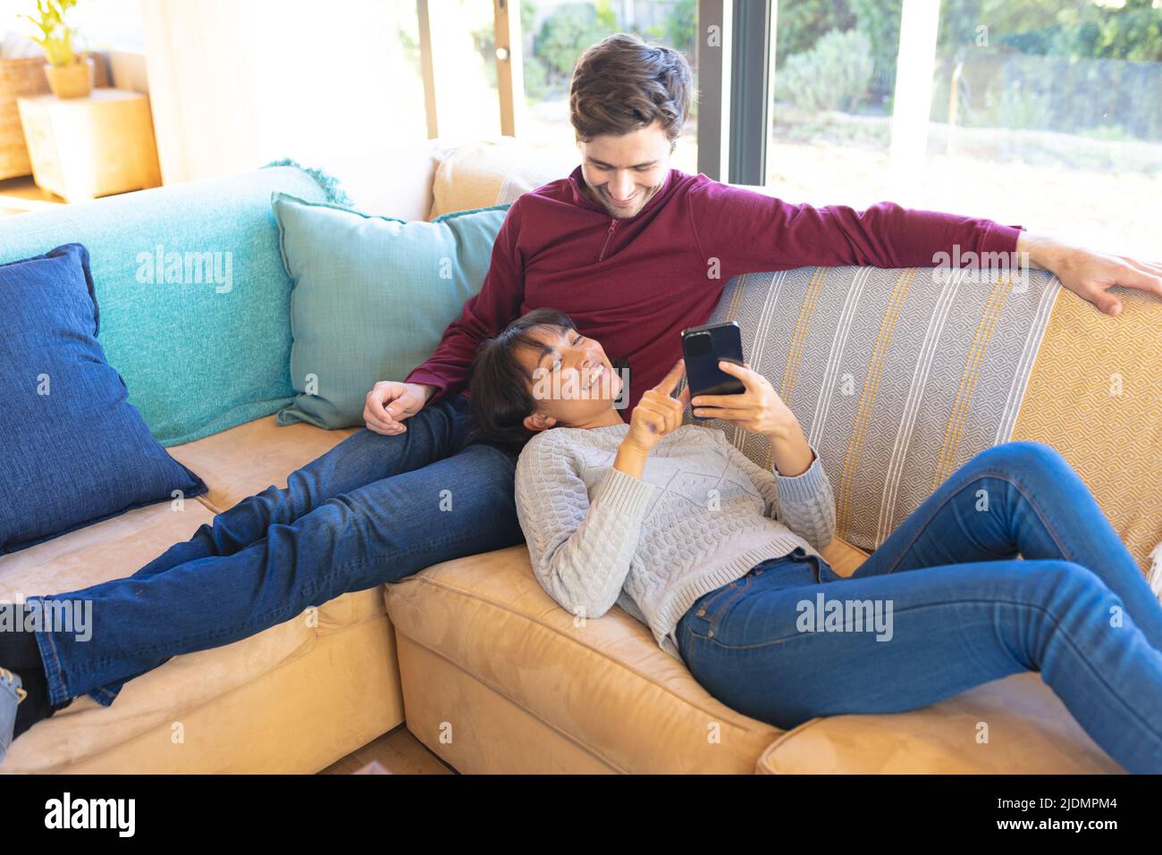 Smiling asian girlfriend using smartphone while lying on caucasian boyfriend's lap on sofa Stock Photo