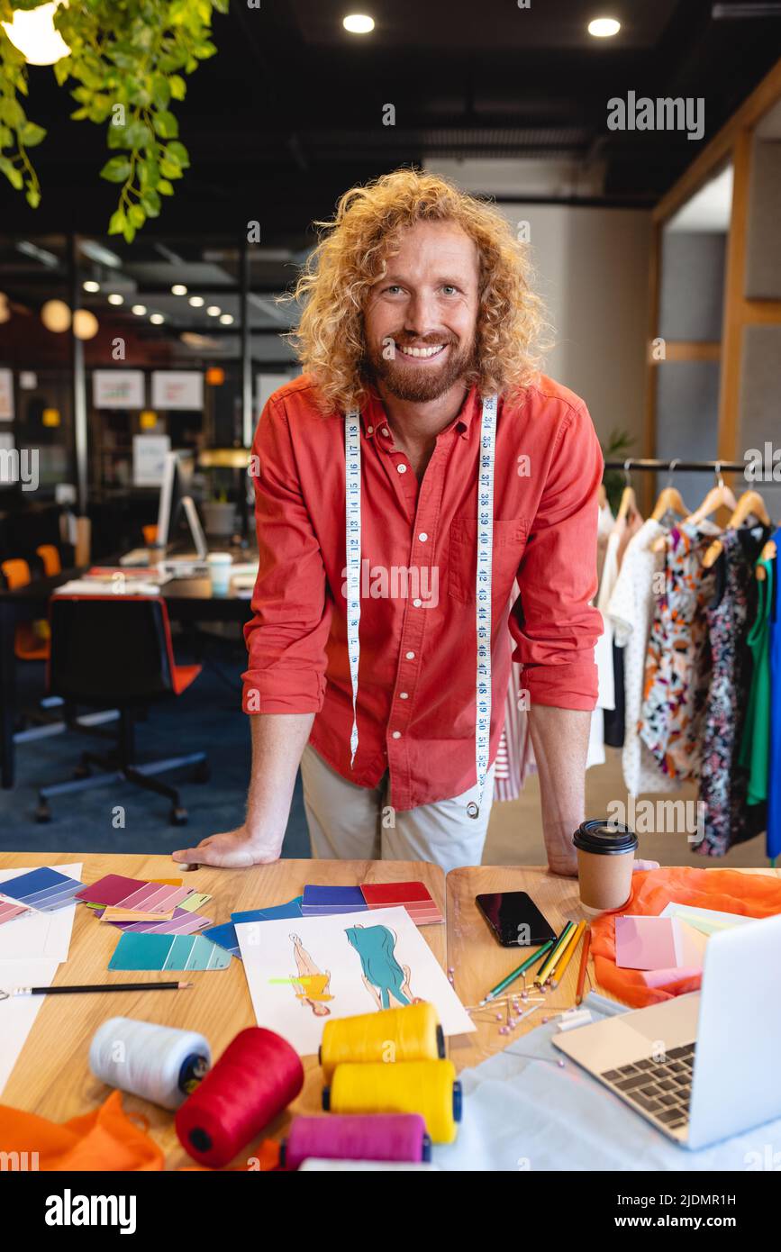 Portrait of smiling caucasian male fashion designer leaning at desk in studio Stock Photo