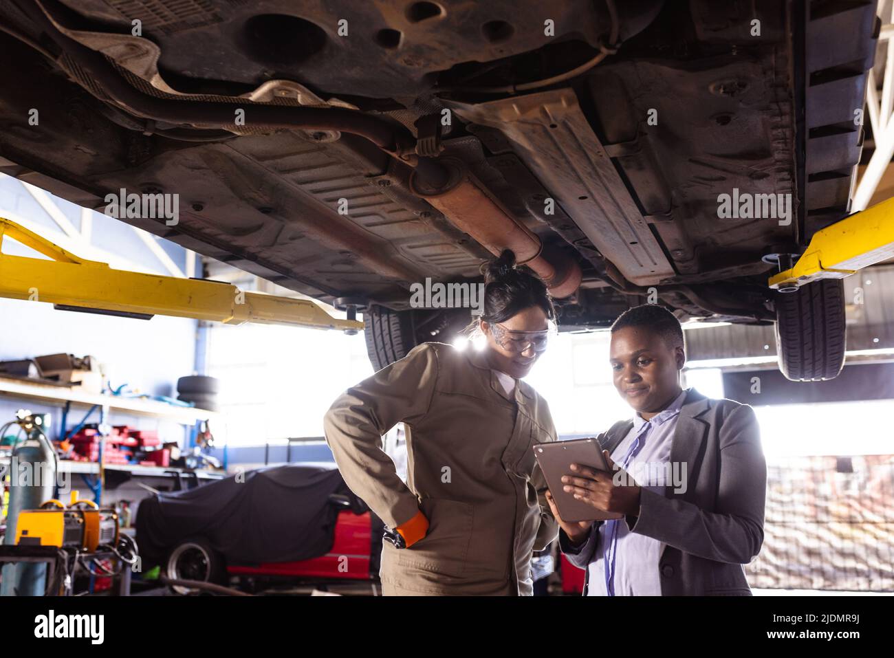 Multiracial mid adult female mechanics repairing car while using digital tablet under car lift Stock Photo