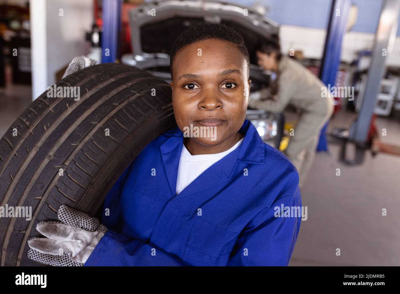 Portrait of confident female mid adult african american mechanic carrying car's tire in workshop Stock Photo