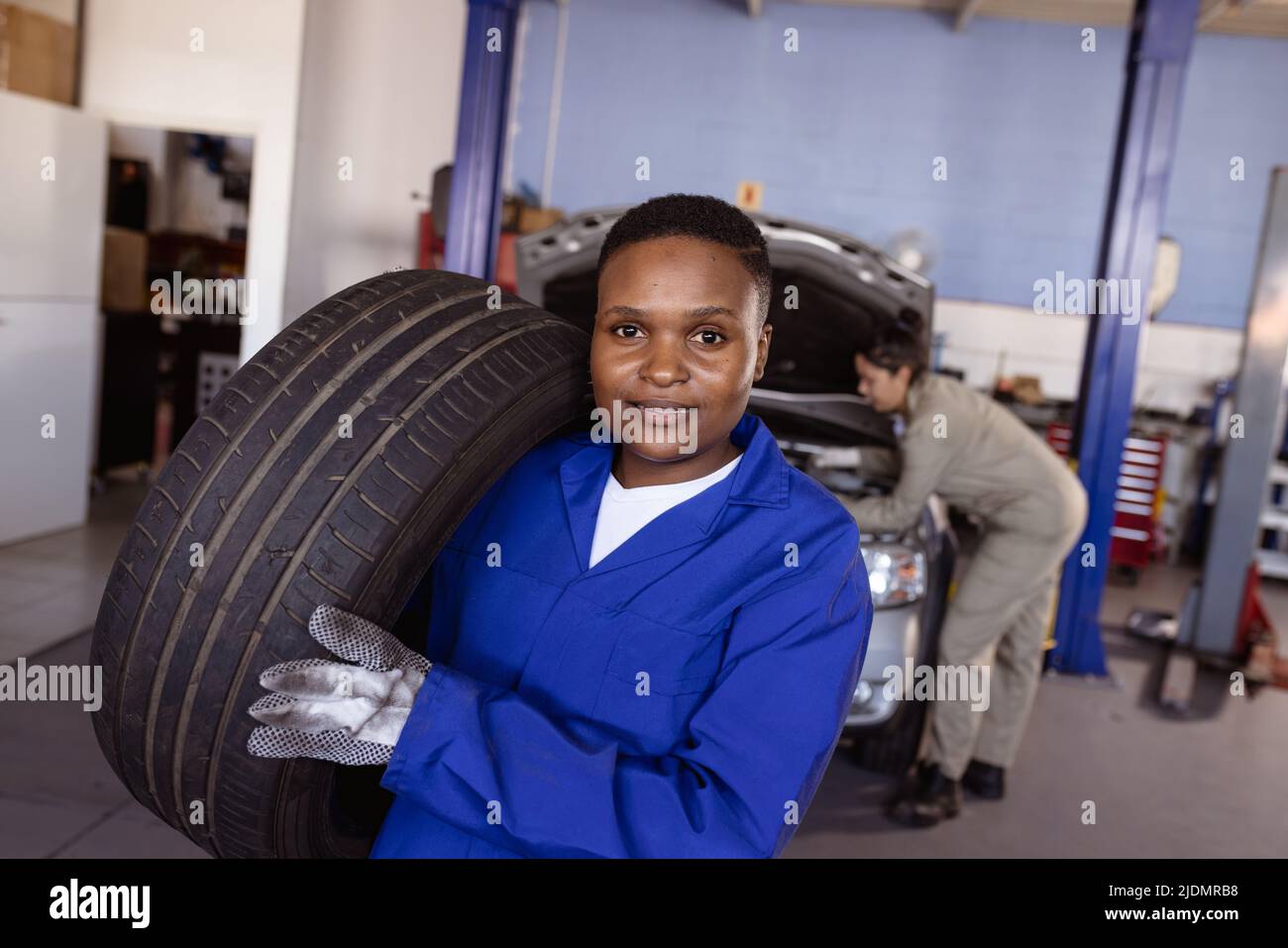 Portrait of confident female mid adult african american welder carrying car's tire in workshop Stock Photo