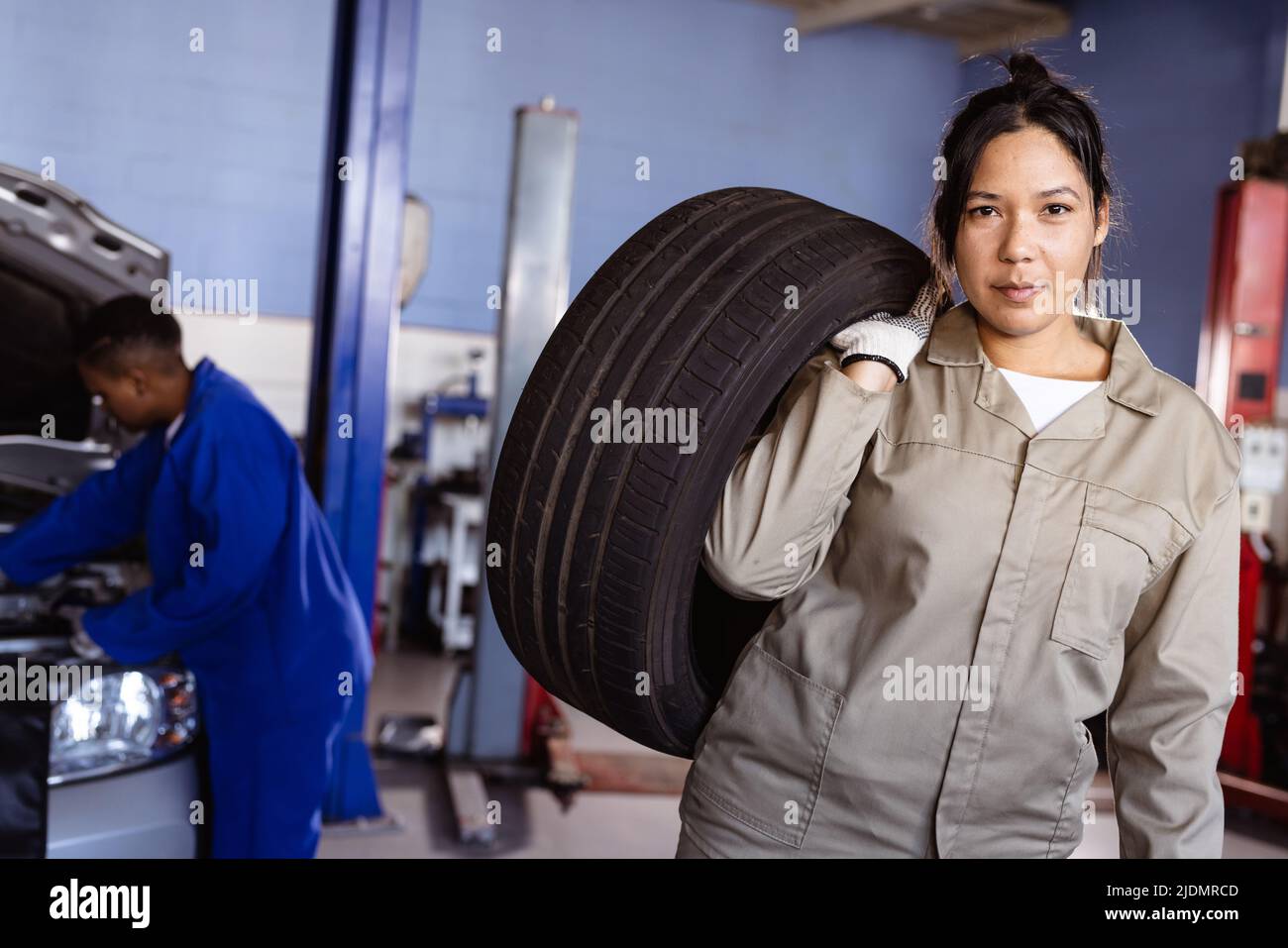 Portrait of confident mid adult asian female mechanic carrying car's tire in workshop, copy space Stock Photo
