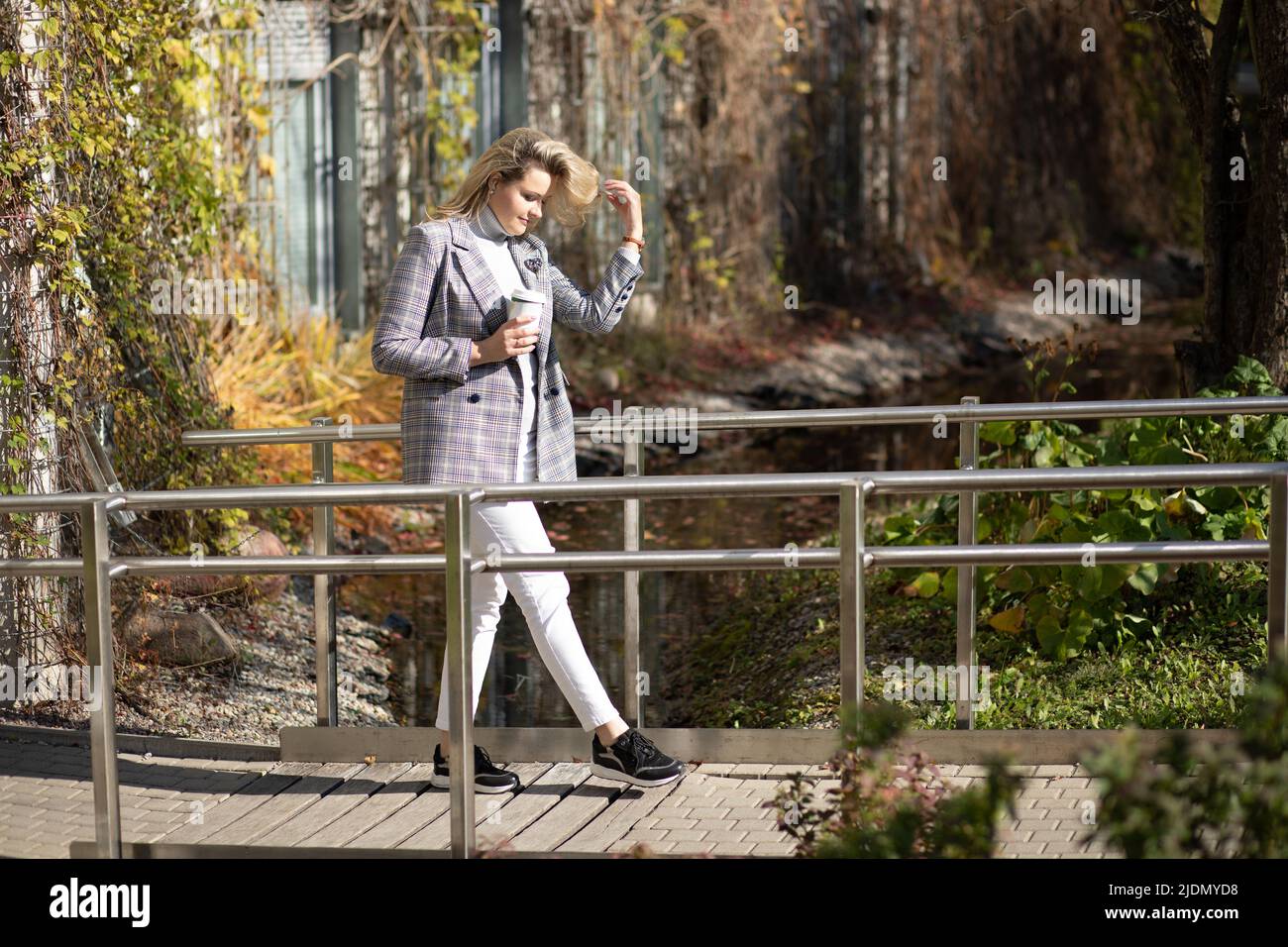 Attractive young woman with long blonde hair in stylish clothes walks along wooden bridge in city park with cup of coffee in her hands . Autumn mood Stock Photo