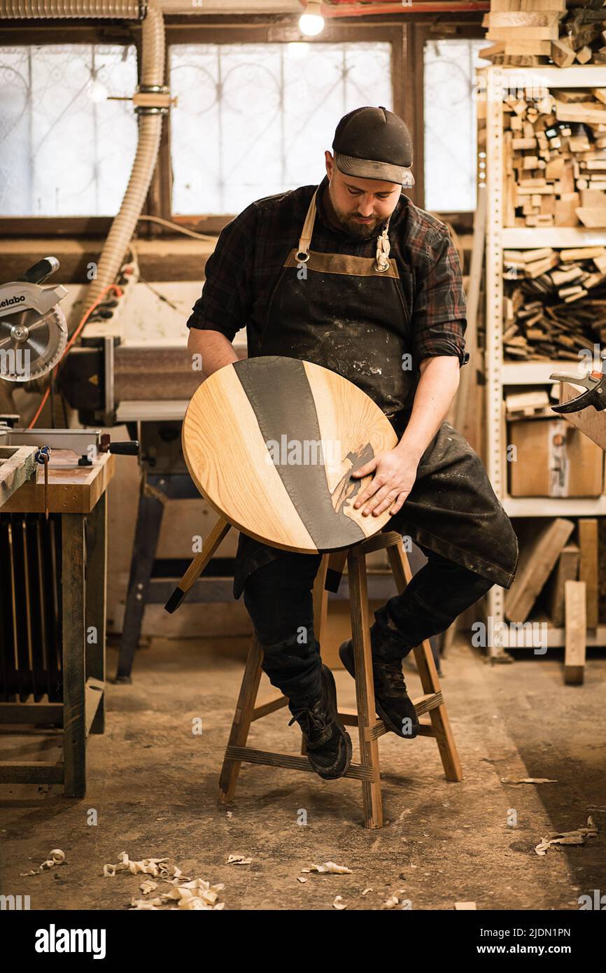 Vertical serious confident bearded carpenter in protective coveralls and cap in store, hold round table. Manual woodwork Stock Photo