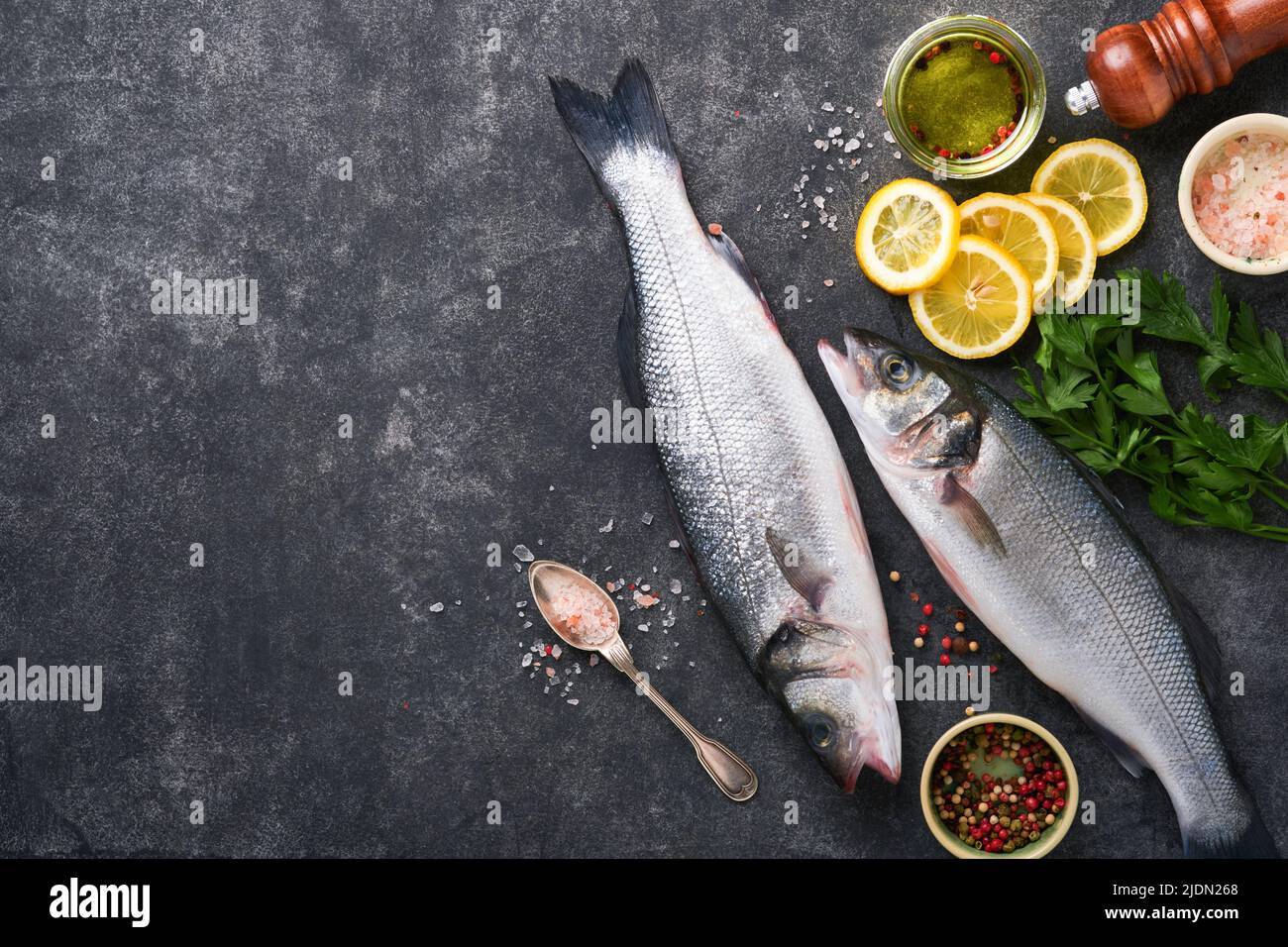 Sea Bass raw. Fresh sea fish bass with salt, pepper, parsley, olive oil and lemon on dark concrete rustic background. Fresh fish ready to cook. Food c Stock Photo