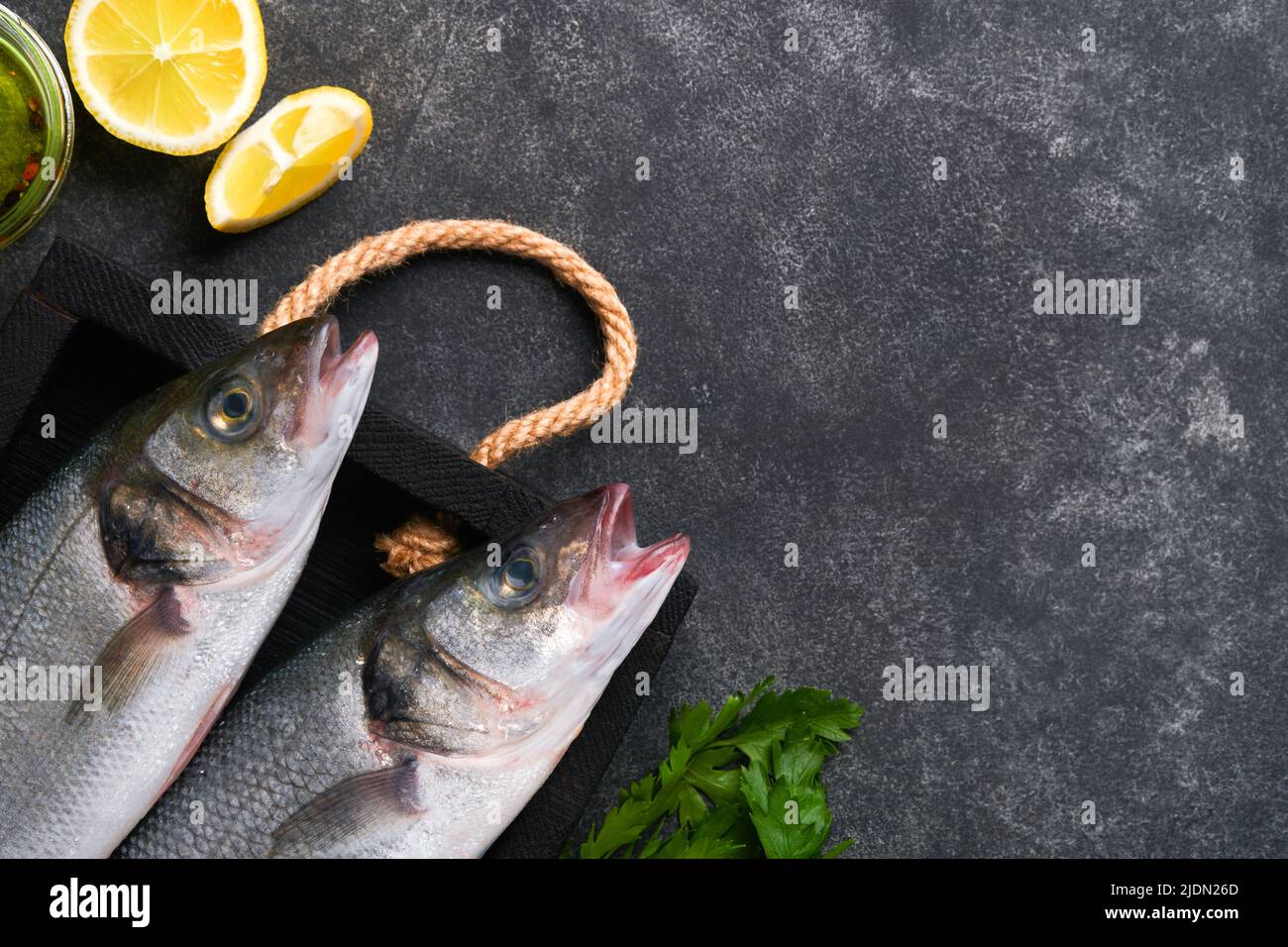 Sea Bass raw. Fresh sea fish bass with salt, pepper, parsley, olive oil and lemon on cutting board on dark concrete rustic background. Food cooking ba Stock Photo