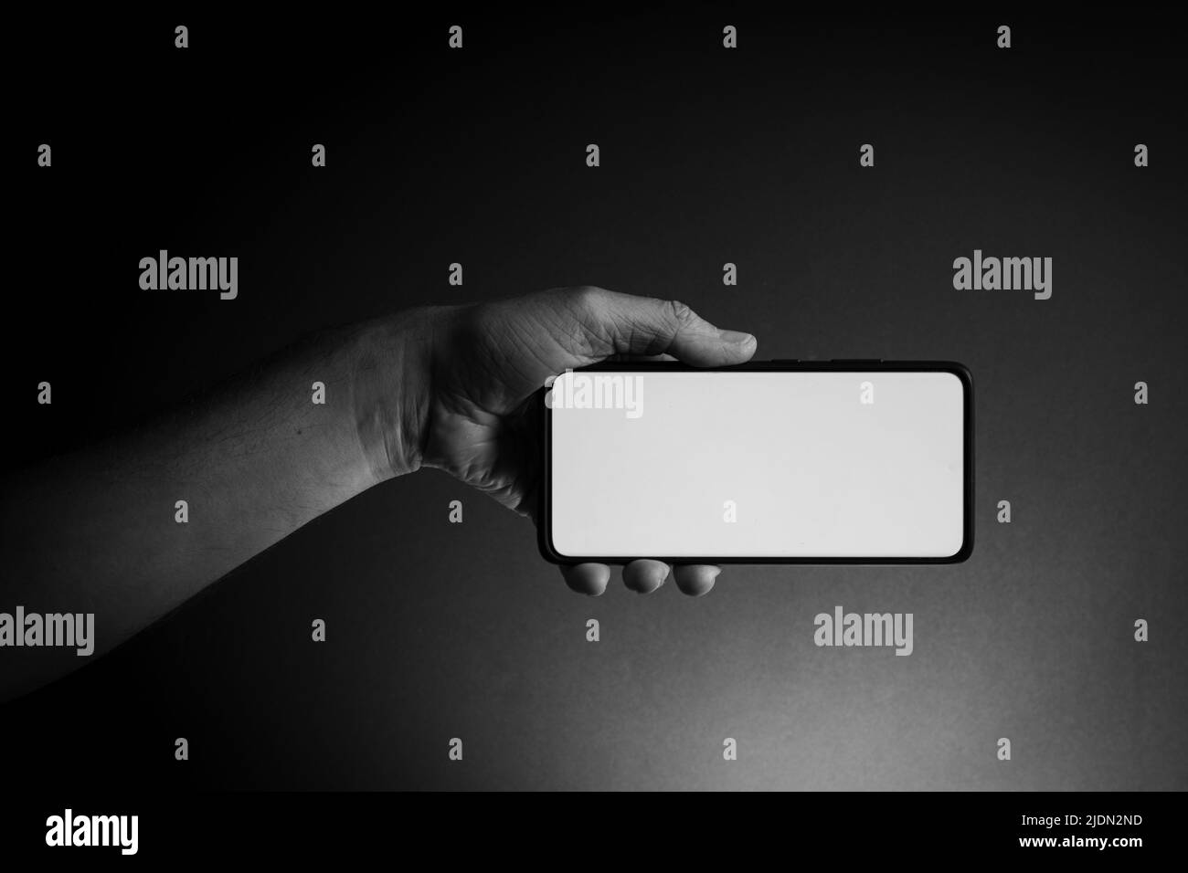 Black and white image of man's hand holding black smartphone horizontally with blank white screen isolated on dark background with dramatic lighting Stock Photo