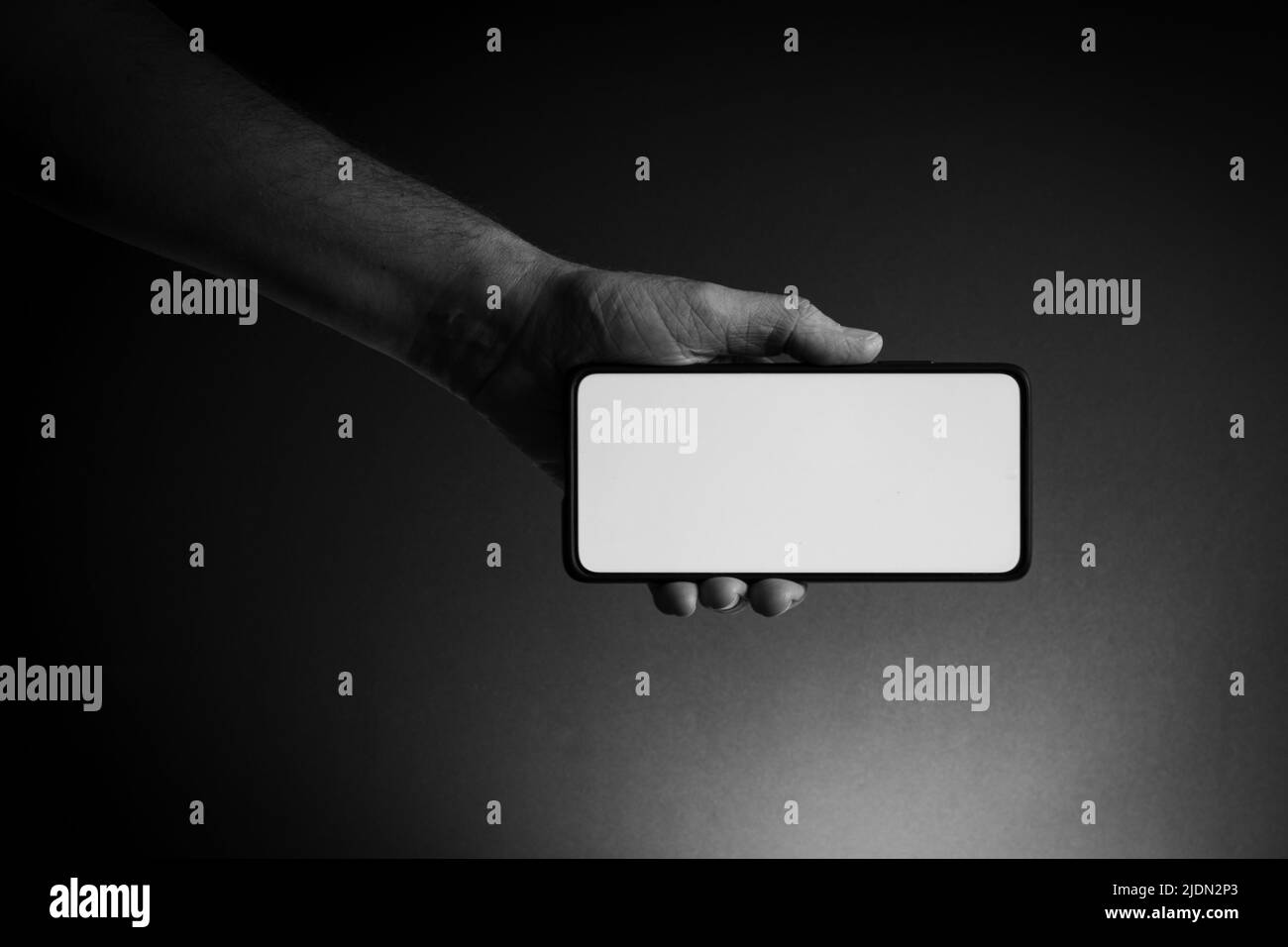 Black and white image of man's hand holding black smartphone horizontally with blank white screen isolated on dark background with dramatic lighting Stock Photo