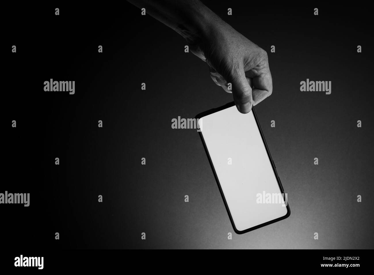 Black and white image of man's hand holding black smartphone with blank white screen isolated on dark background with dramatic lighting and copy space Stock Photo