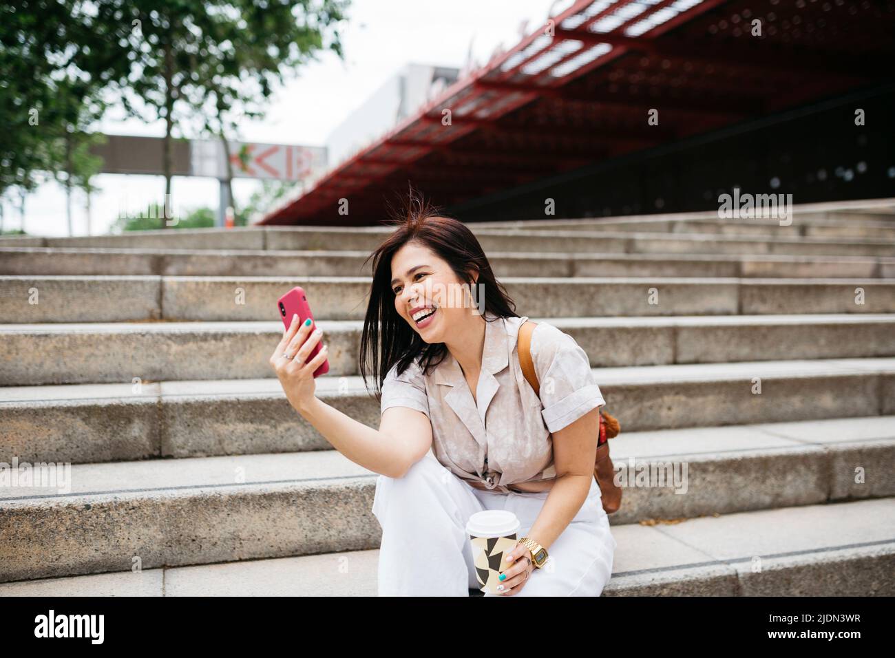 Young brunette woman sitting on stairs with a coffee to go and taking a selfie Stock Photo