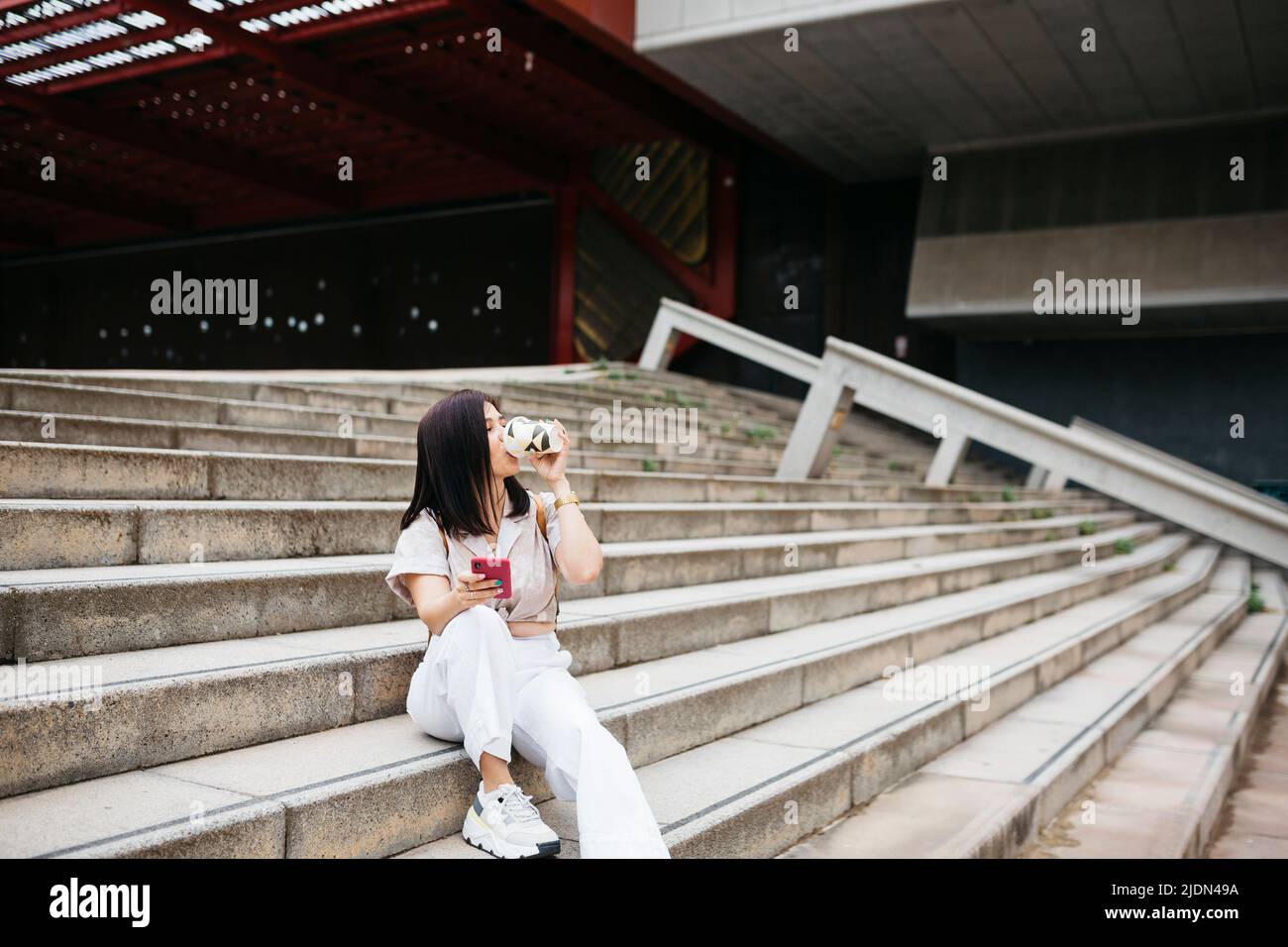 Young brunette woman sitting on stairs drinking coffee and using a phone Stock Photo